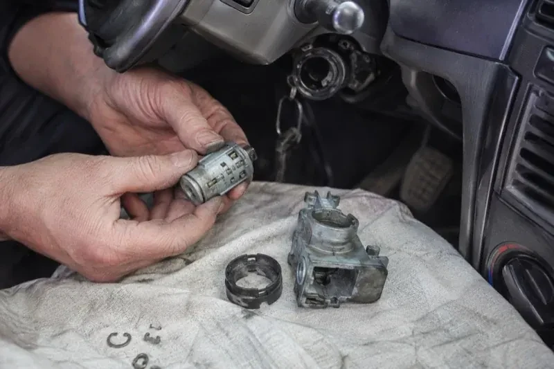 Hands holding a removed ignition cylinder beside a car steering column, with the housing and small parts on a cloth. — Ignition Lock Repair, Innisfil